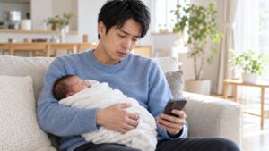明るいリビングで新生児を抱き、スマートフォンを見て困った表情をする男性